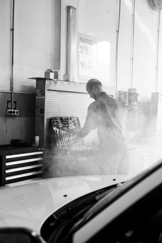 Mechanic in a garage performing vehicle maintenance amidst tools and equipment in a smokey atmosphere.